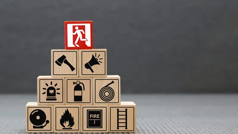 Cubos de madera apilados en pirámide con íconos de protección contra incendios (extintor, manguera, alarma, hacha, letrero FIRE), coronados por un cubo rojo con el pictograma de una salida de emergencia