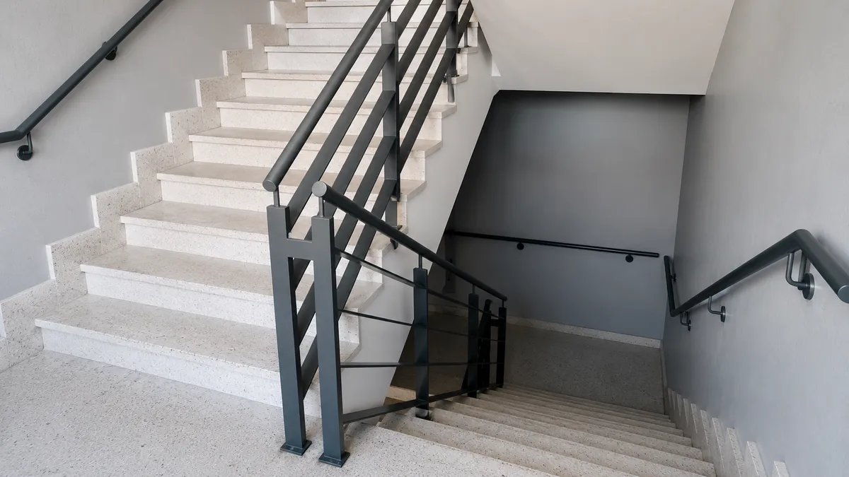 Escalera interior de edificio vista desde el nivel superior, con dos tramos de escalones de concreto claro que giran en torno a un descanso, pasamanos metálicos gris oscuro y barandal de balaustres verticales, y paredes gris claro