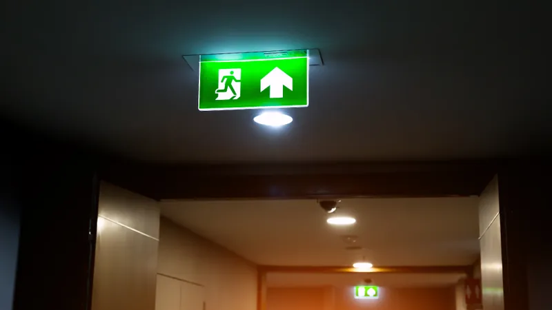Vista de un pasillo de oficina con un flujo de personas caminando de forma ordenada hacia una puerta de salida de emergencia iluminada, con señalización verde de ruta de evacuación en el techo