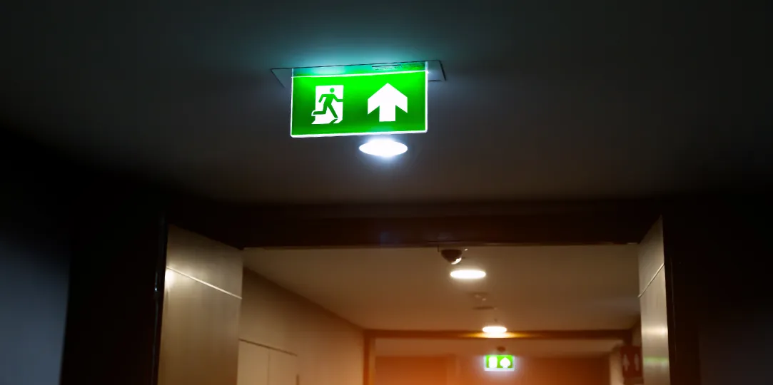 Vista de un pasillo de oficina con un flujo de personas caminando de forma ordenada hacia una puerta de salida de emergencia iluminada, con señalización verde de ruta de evacuación en el techo