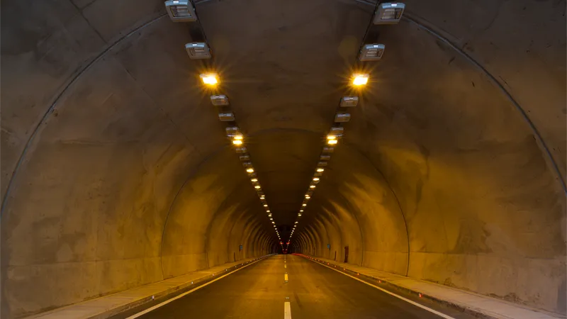 Vista interior de un túnel vehicular iluminado con luces LED en el techo y ductos de ventilación metálicos a lo largo de las paredes, con el trazado de la carretera perdiéndose en la distancia