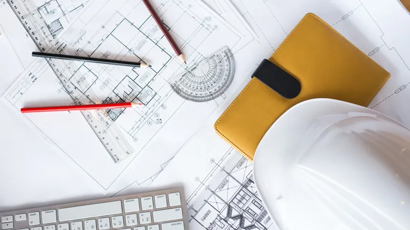 Primer plano de planos arquitectónicos de un centro comercial extendidos sobre una mesa de trabajo, junto a un casco de ingeniero, un lápiz y una calculadora